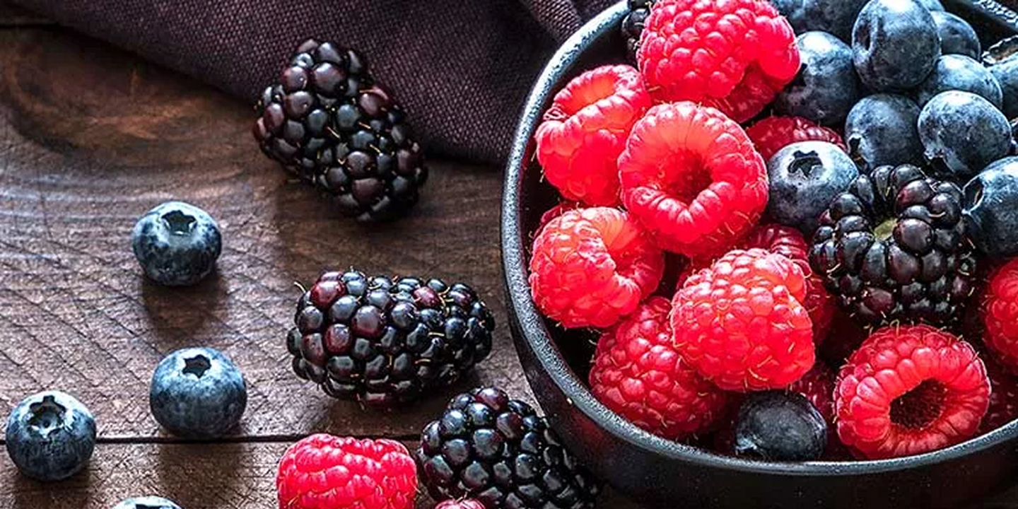 A close-up of a dark bowl filled with raspberries, blueberries, and blackberries on a wooden surface.