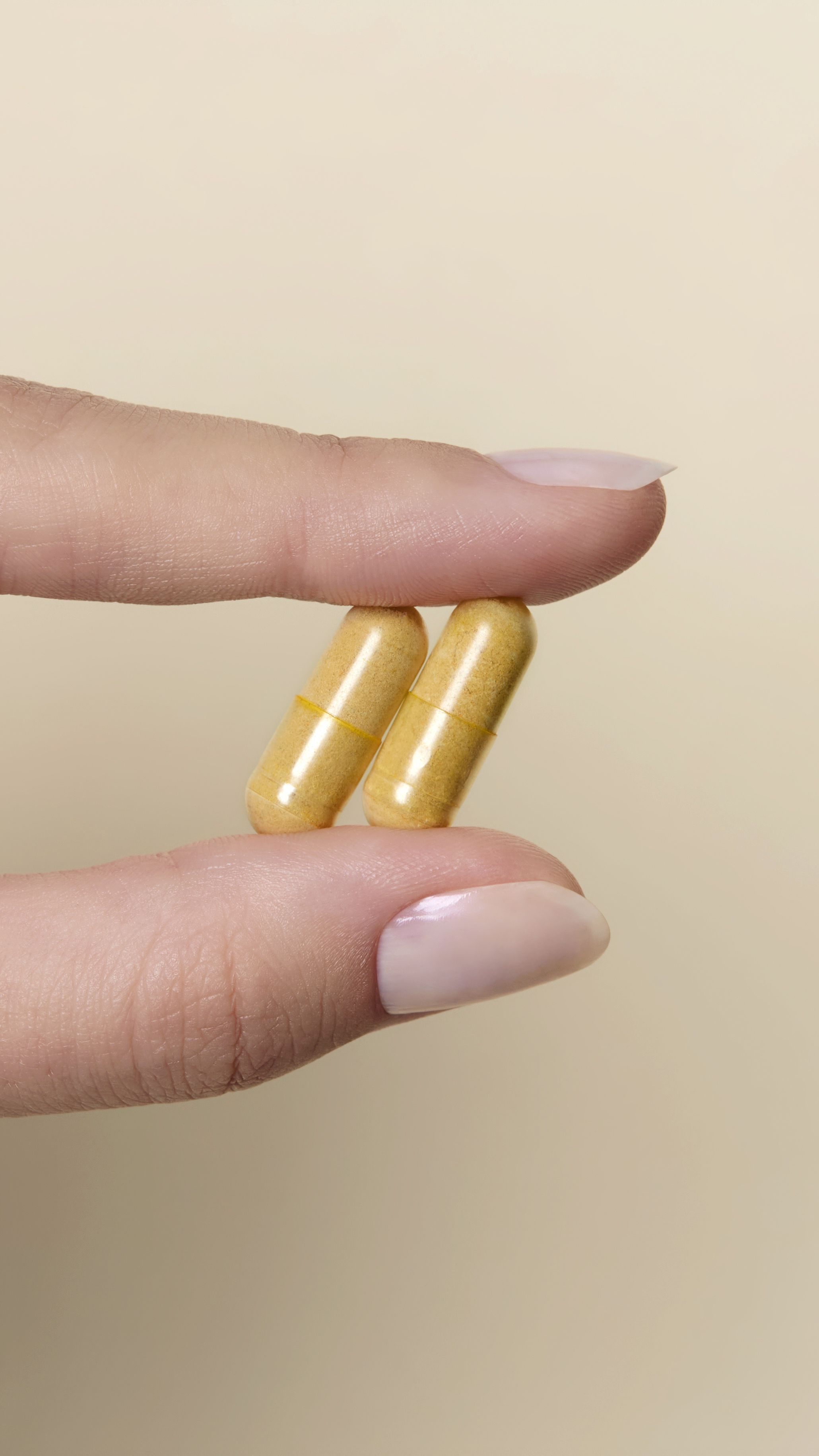 A person's fingers with a light pink manicure holding two yellow capsules against a beige background.