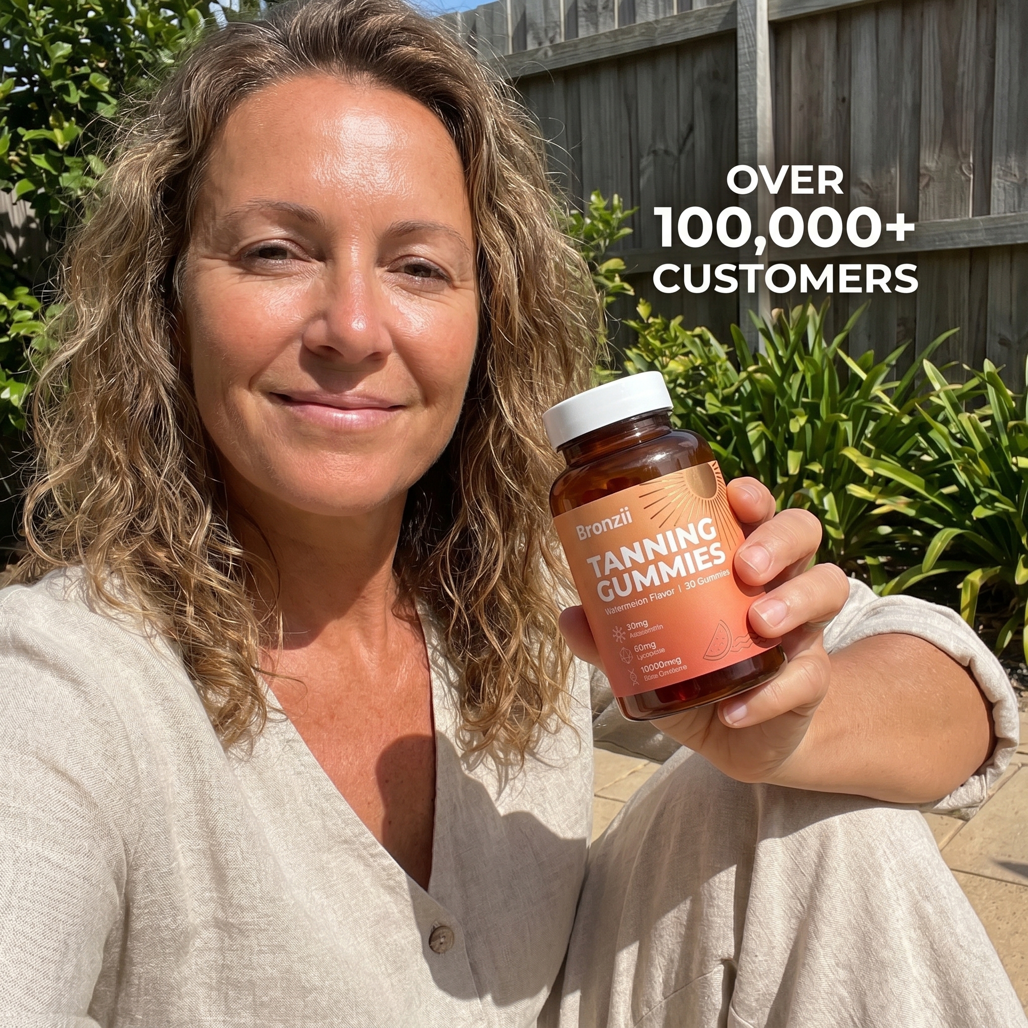 A woman with wavy hair smiles while holding a bottle of Bronzii Tanning Gummies outdoors in the sun.