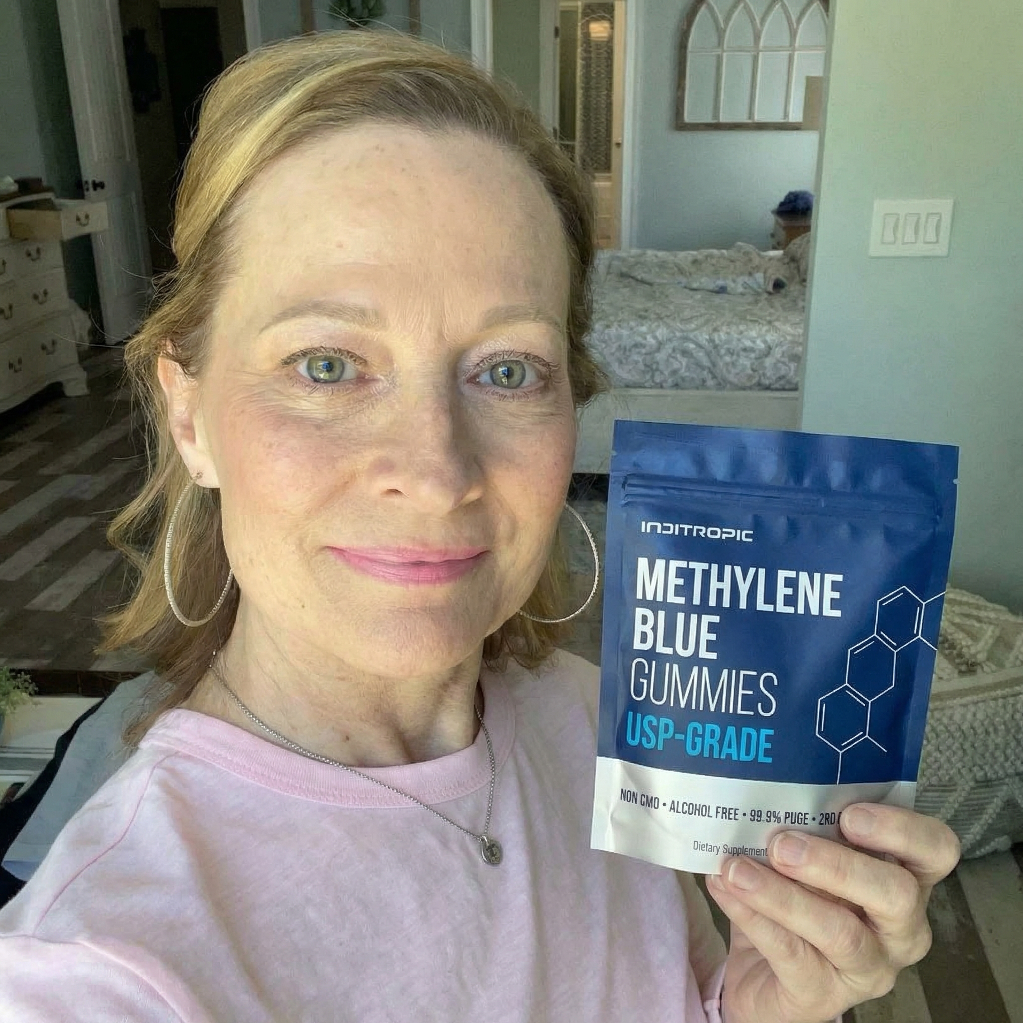 A smiling woman holds up a blue pouch of Inditropic Methylene Blue Gummies in her bedroom.