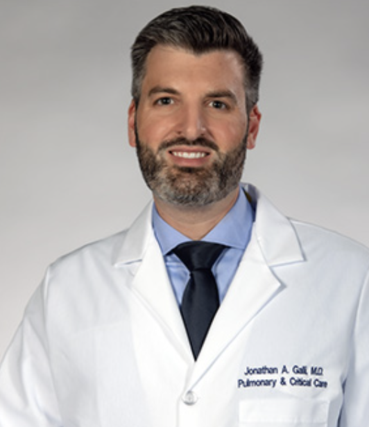 Person in a white coat with a tie, smiling.