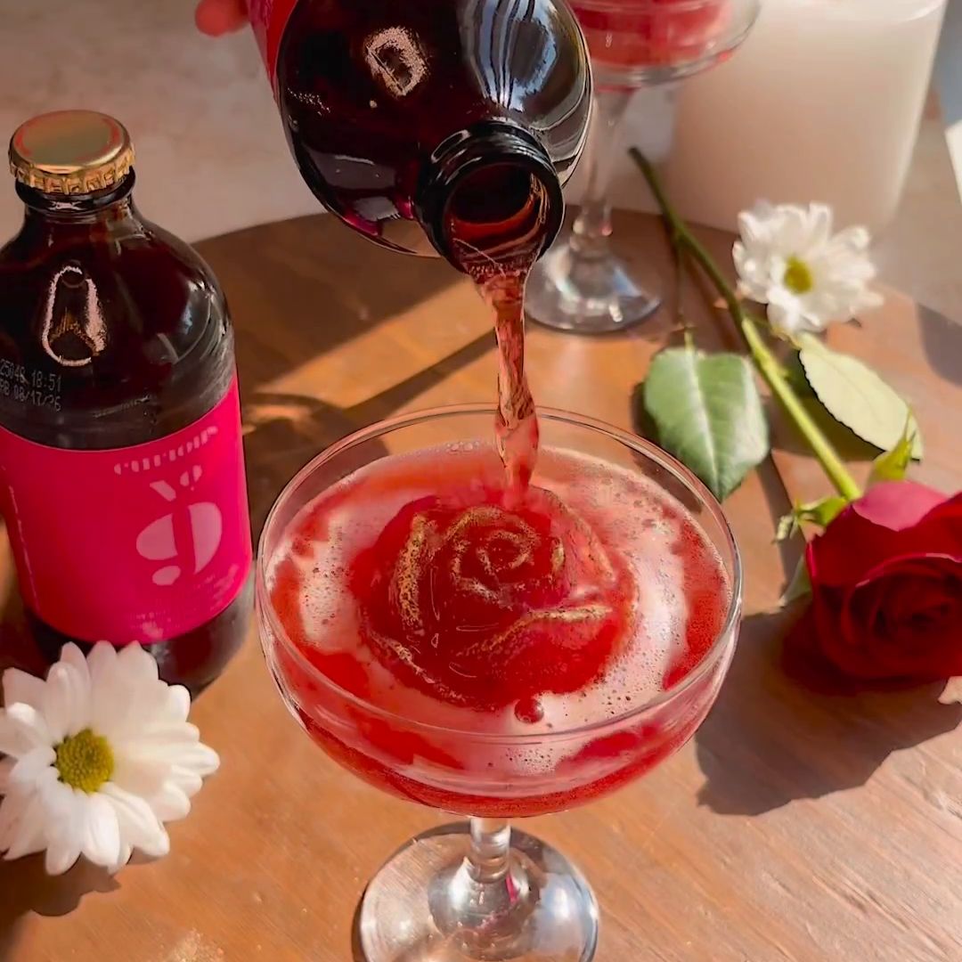 A red beverage is poured from a bottle into a coupe glass containing a rose-shaped ice cube.