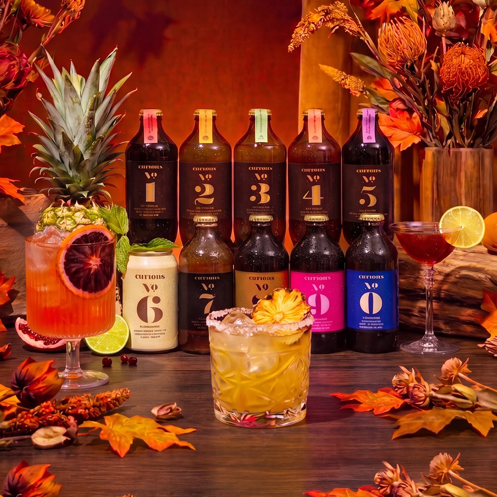 An autumnal display of Curious brand bottled drinks and three garnished cocktails on a wooden table.
