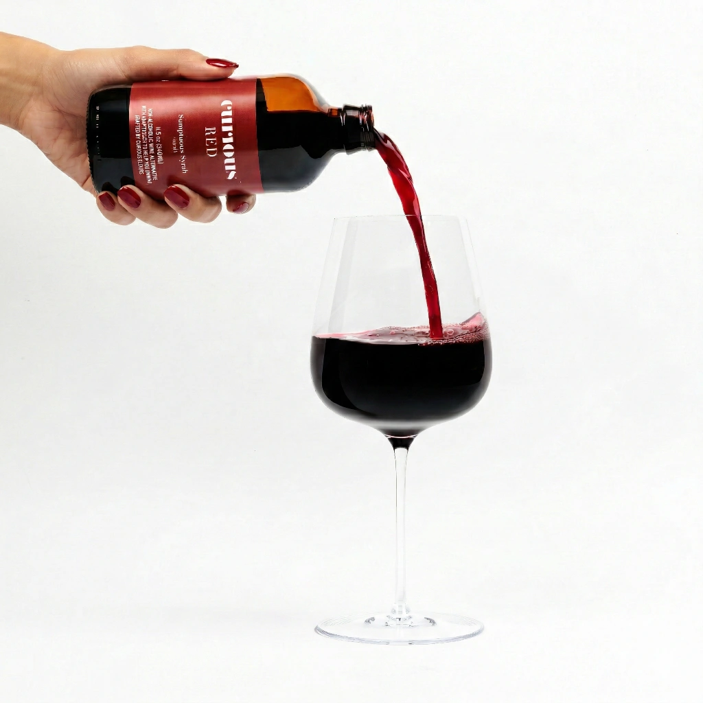 A hand pours a red liquid from a bottle labeled 'Curious RED' into a large wine glass.