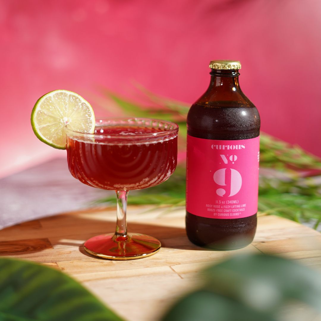 A bottle of Curious No. 9 sits next to a poured cocktail in a glass with a lime garnish.