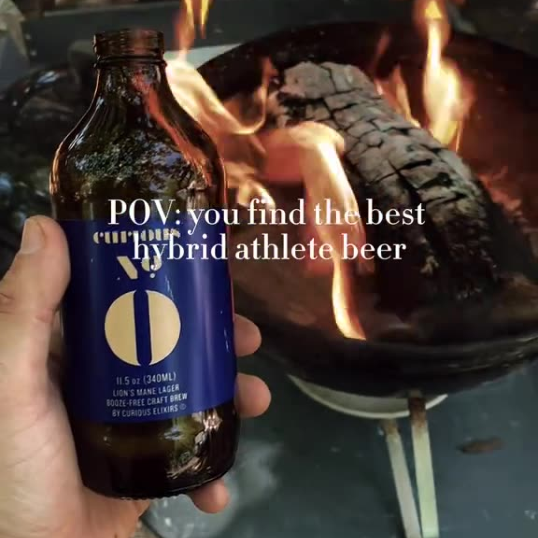 Hand holding a beer bottle in front of a fire pit with burning wood.