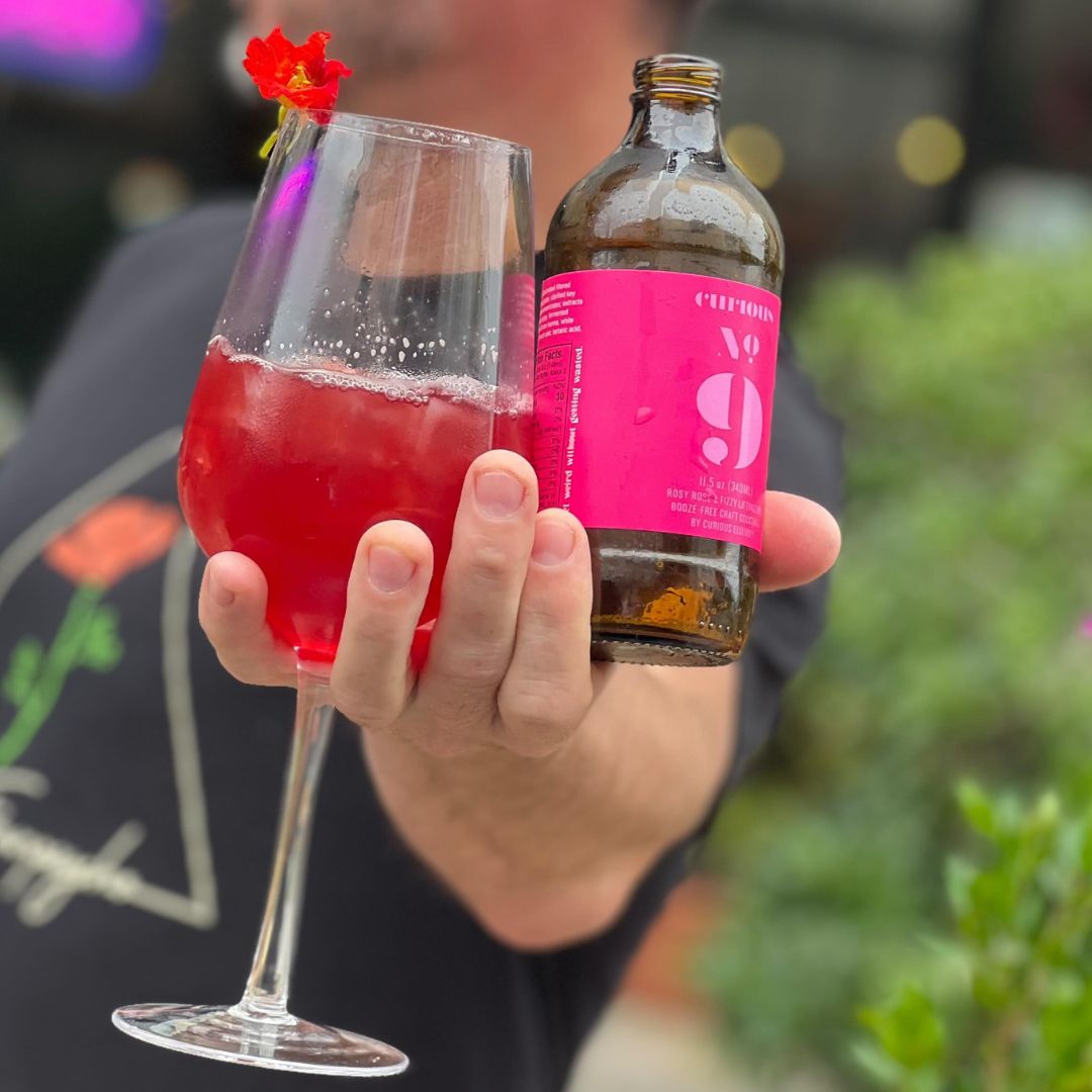A person holds a glass of a pink beverage with a flower garnish and a bottle of Curious No. 9.