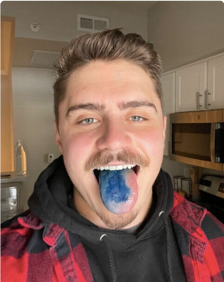 Person with blue tongue, wearing a red and black plaid shirt, in a kitchen.