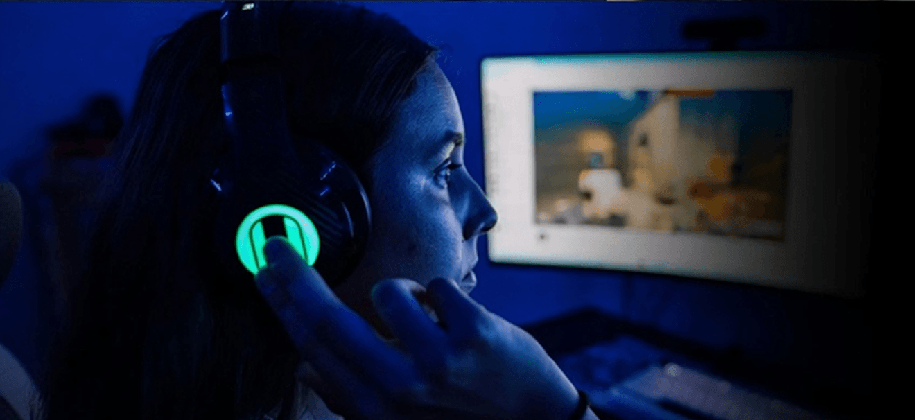 A person wearing glowing headphones looks at a computer screen in a dark, blue-lit room.