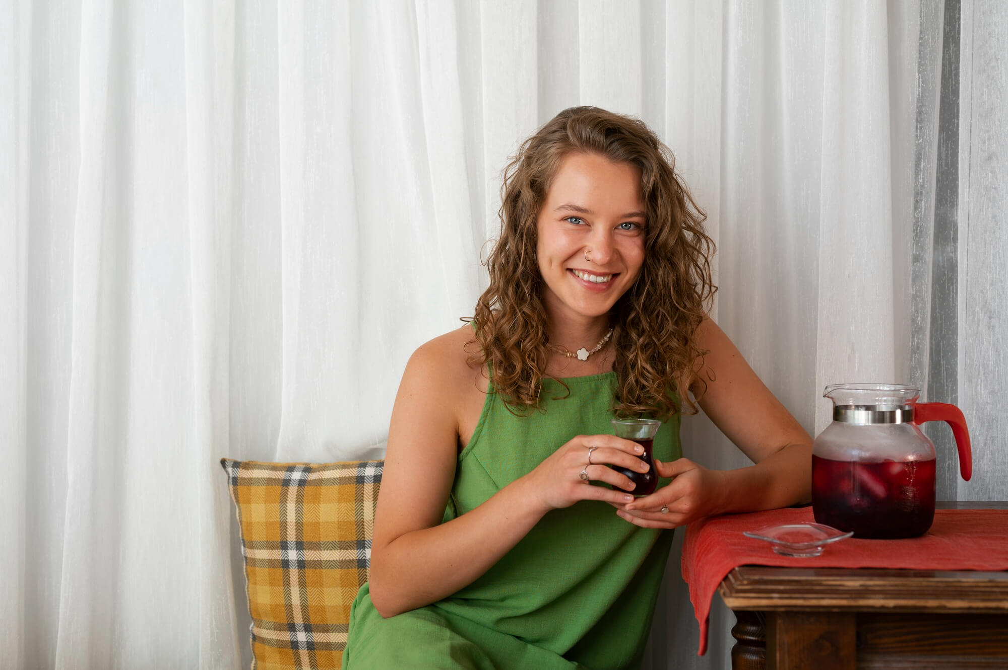 A smiling woman in a green dress sits at a table holding a glass of a red beverage.
