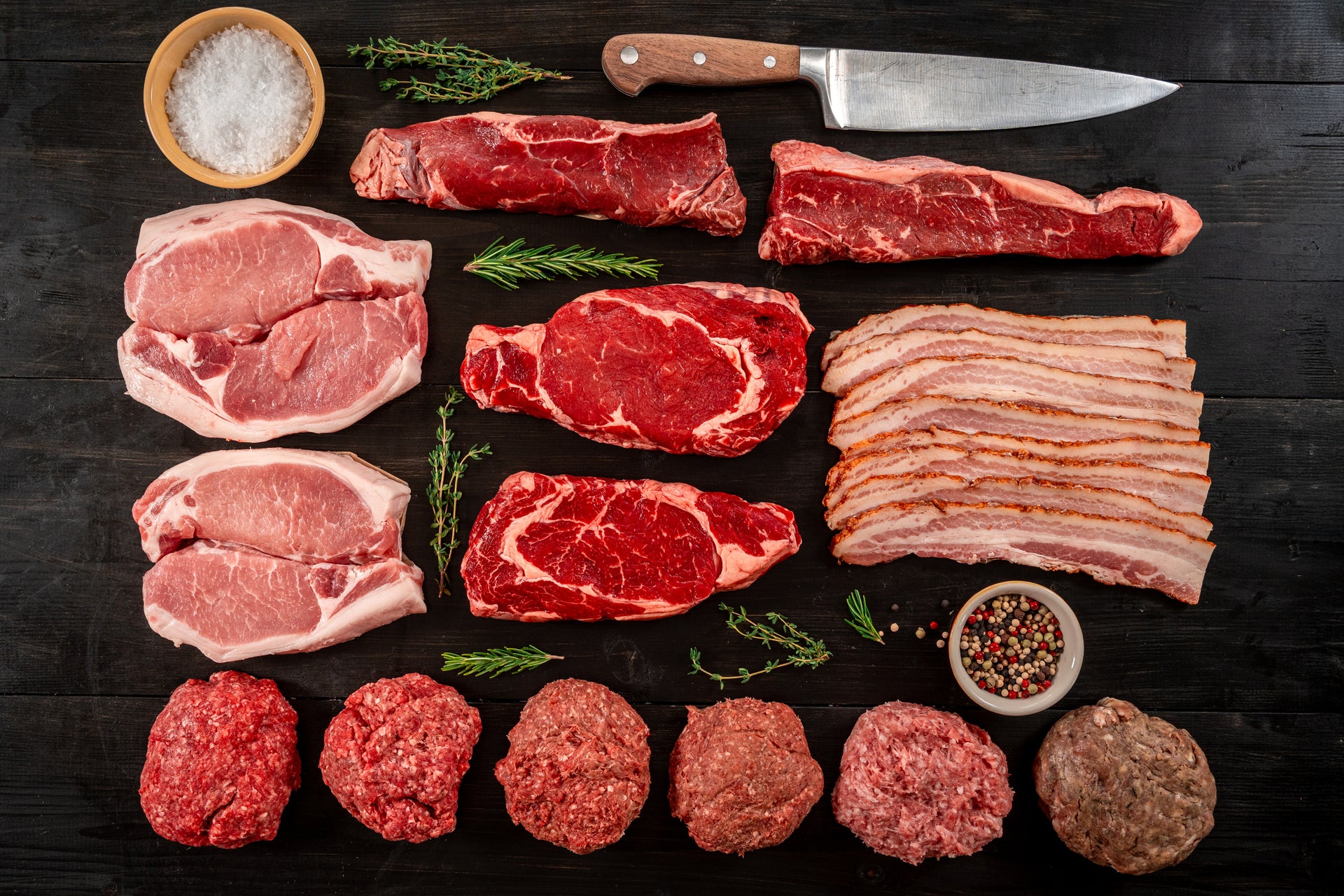 Various cuts of raw meat displayed with herbs, knife, and bowl of salt and peppercorns.
