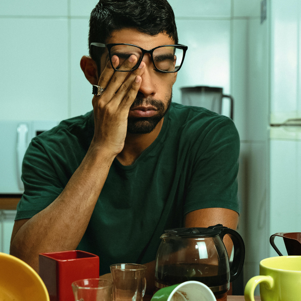 Man with glasses holding face, surrounded by coffee cups.