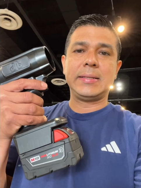 A man in a blue t-shirt holds a black cordless power tool up to the camera.