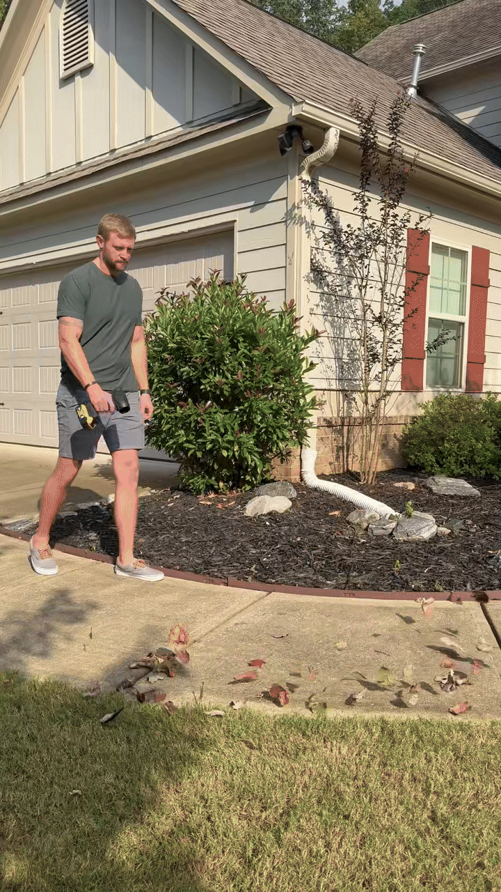 Man using a leaf blower on a driveway next to a garden.