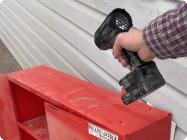 A person holds a black cordless blower over a red metal cabinet next to a wall with siding.