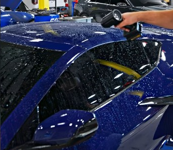 A person holding a tool over the wet window of a blue car in an auto shop.