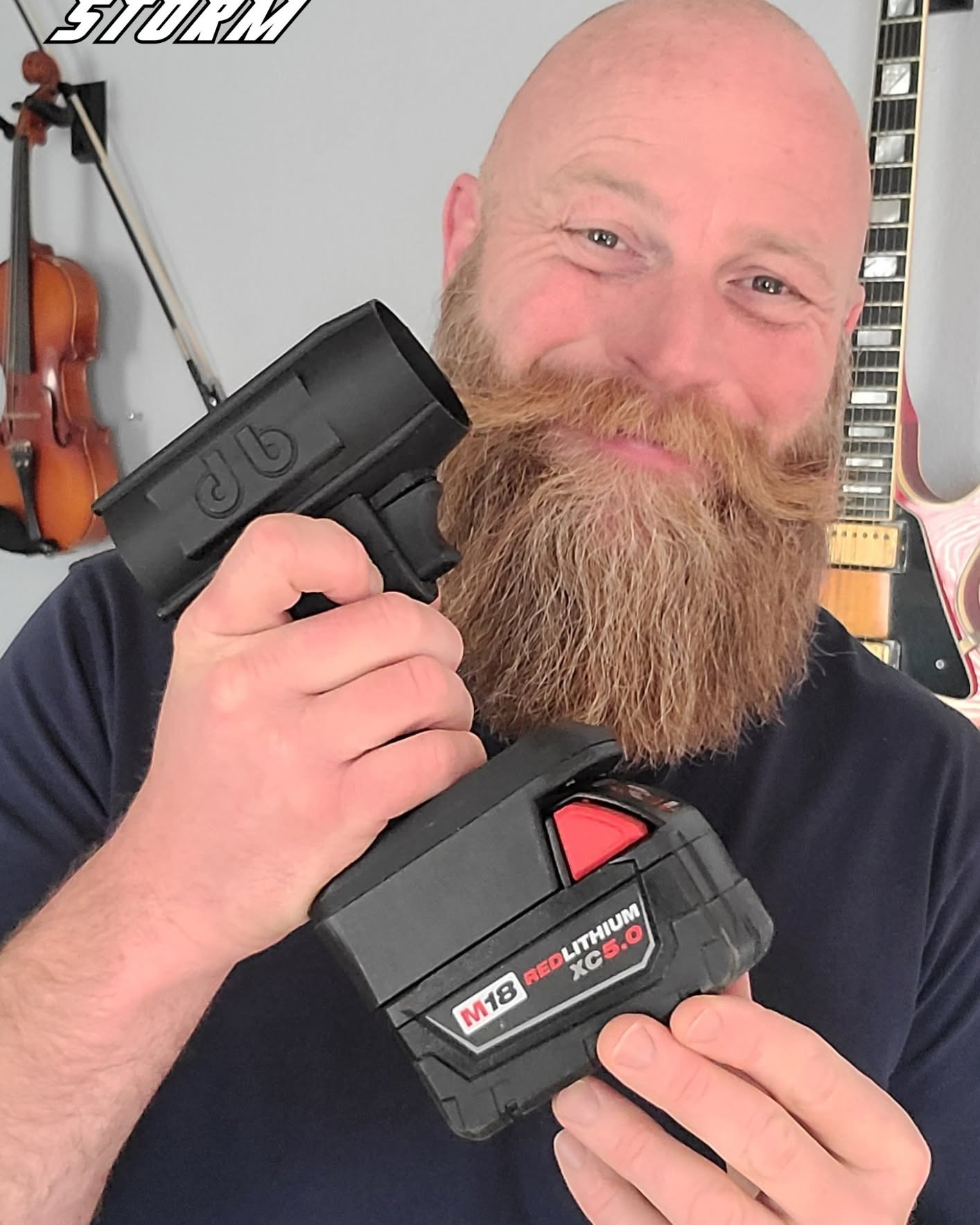 A smiling bald man with a large beard holds a black tool part and an M18 battery.