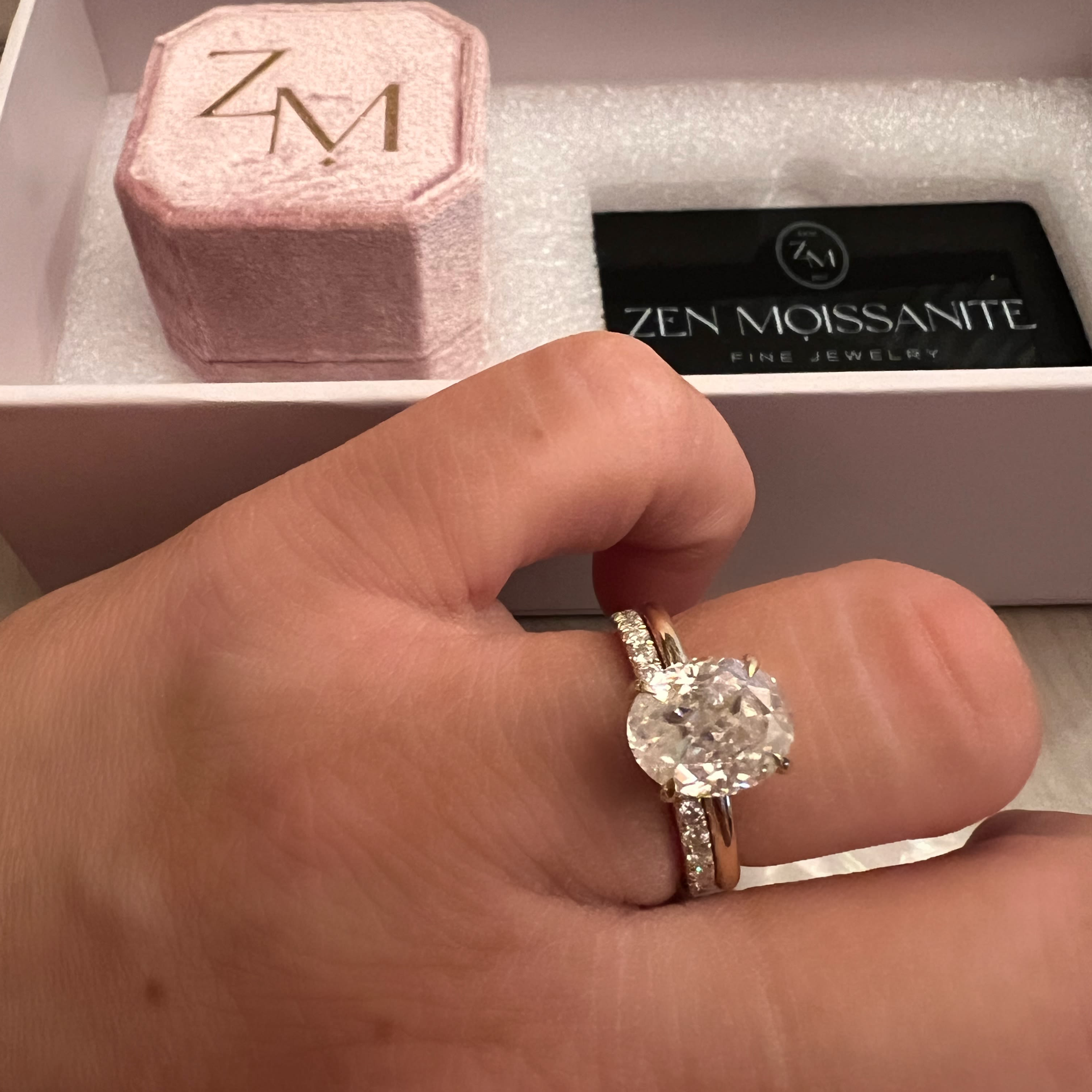 A hand wearing a gold oval engagement ring and a diamond band, with a pink ring box behind it.