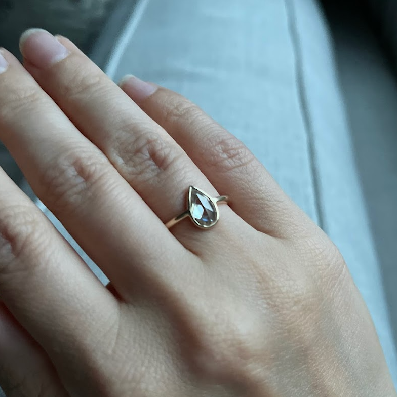 A close-up of a hand wearing a delicate ring with a pear-shaped, bezel-set stone.