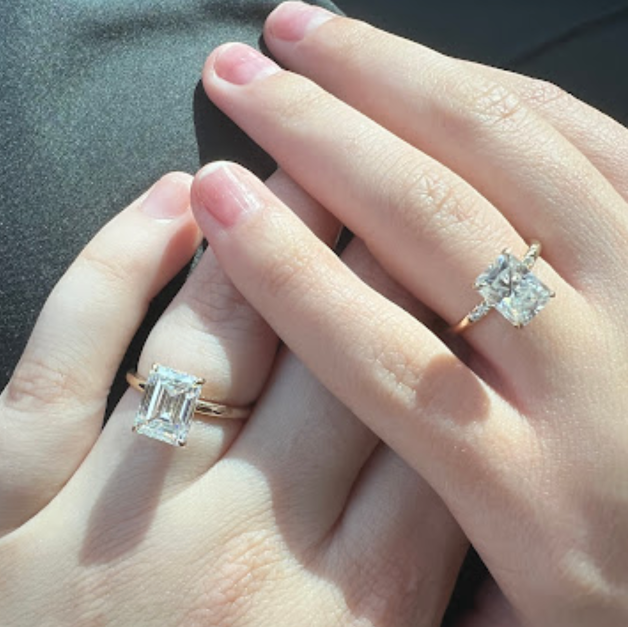 A close-up of two hands intertwined, each wearing a different style of large diamond ring.