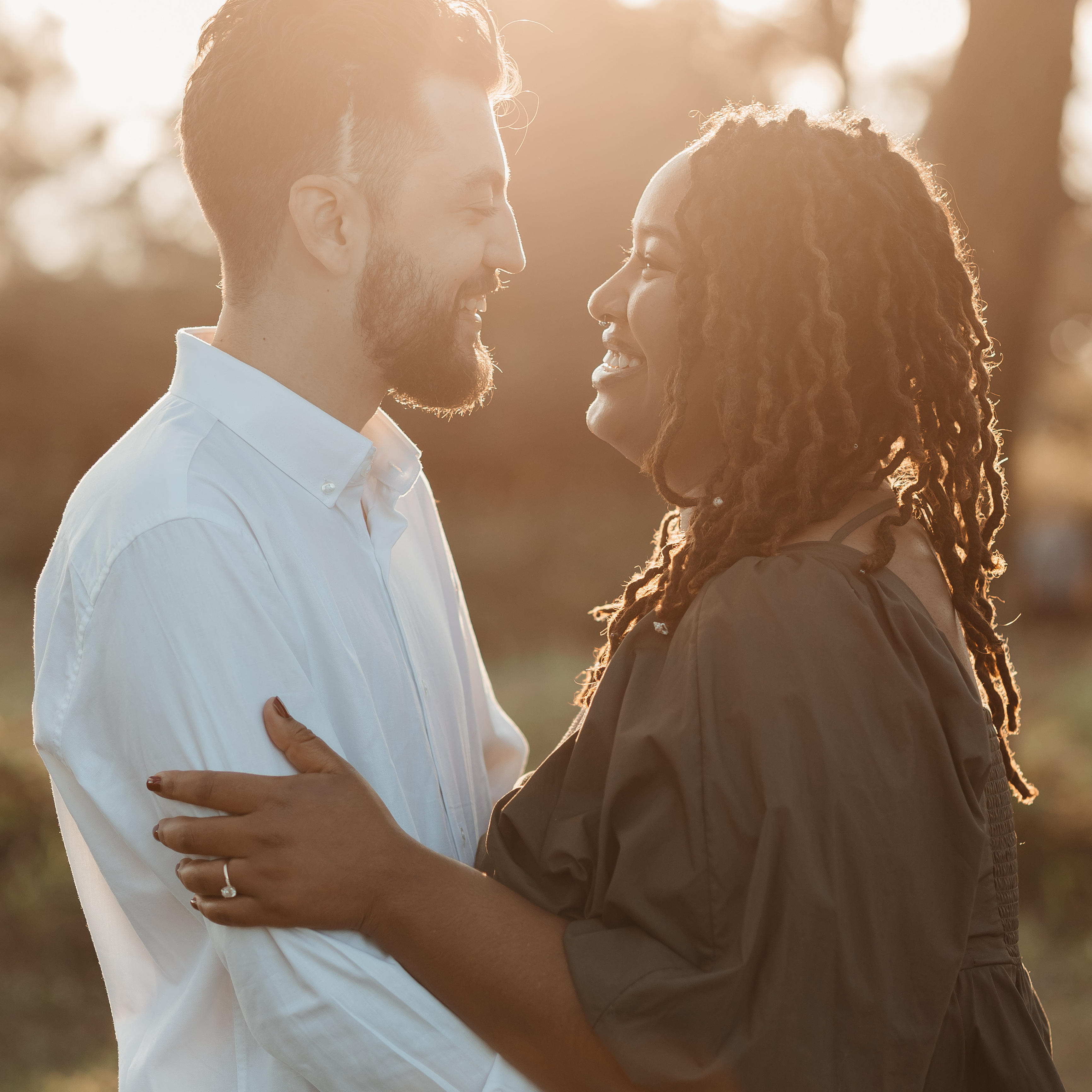 A man and a woman embrace and smile at each other outdoors, backlit by golden sunlight.