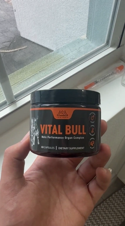 Hand holding a jar labeled 'Vital Bull' dietary supplement near a window.