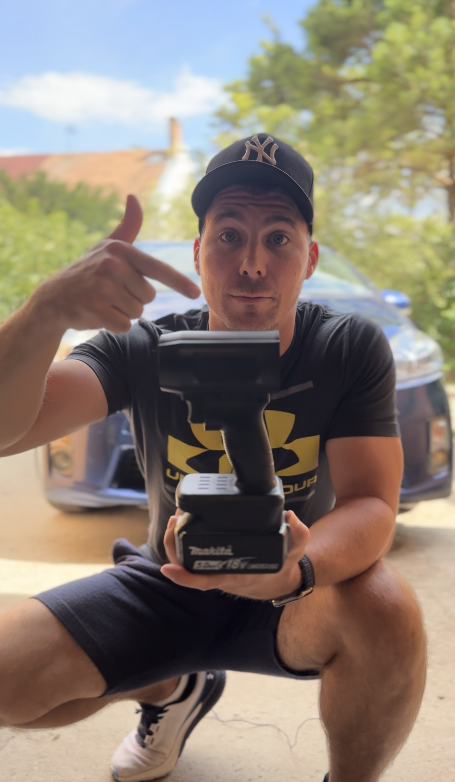 Person holding a Makita power tool battery, pointing at it, with a car in the background.