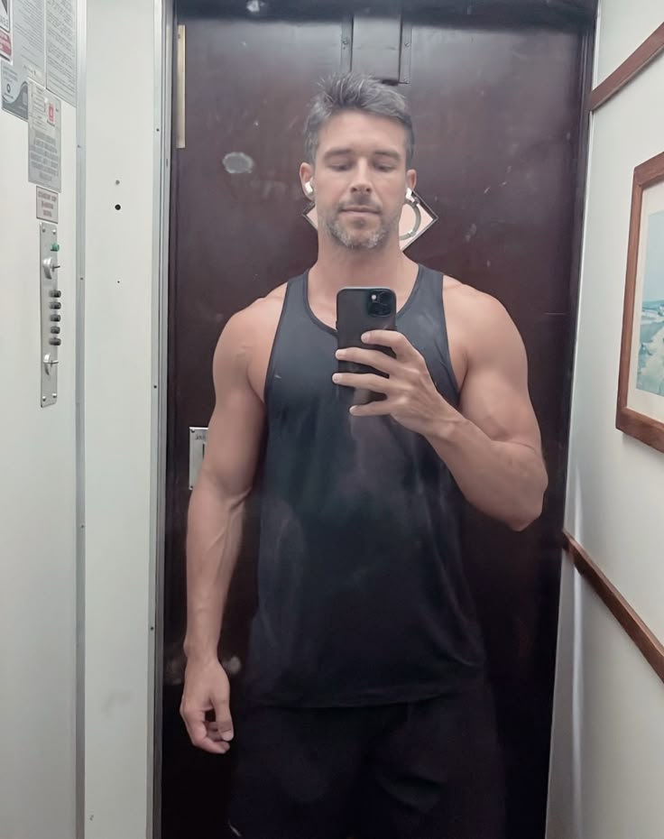 Man taking a mirror selfie in an elevator, wearing a black tank top.