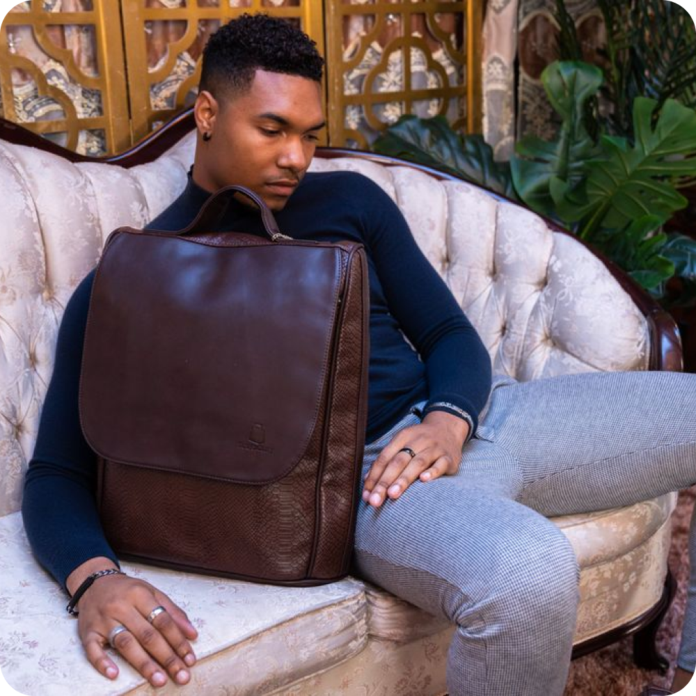 A man sits on a vintage-style couch, looking down at a brown leather backpack placed next to him.