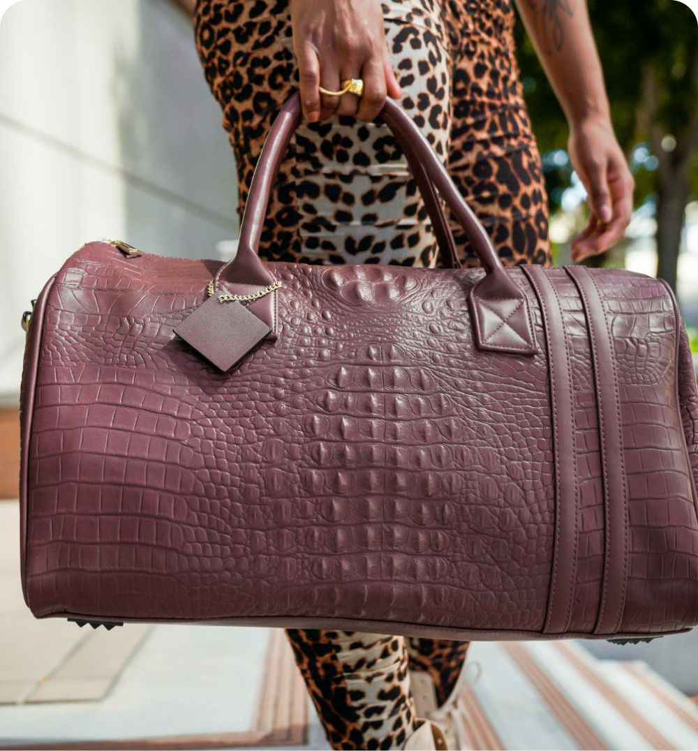A person in leopard print pants holding a large, burgundy, crocodile-textured travel bag.