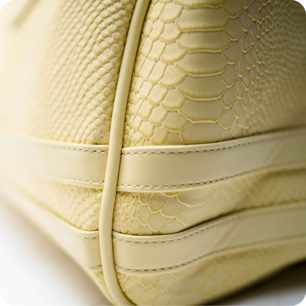 A close-up of the corner of a pale yellow, snakeskin-textured leather bag with stitching detail.