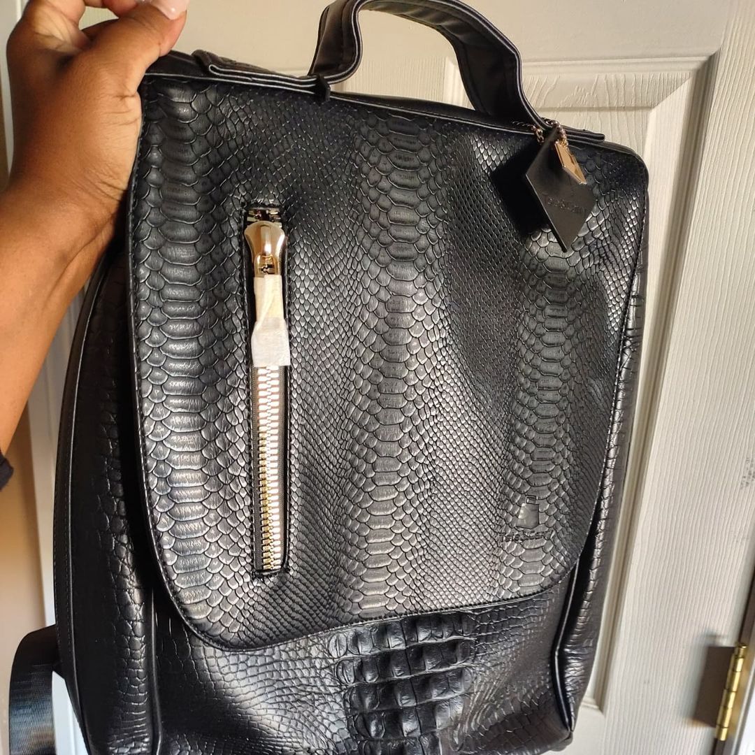A hand holds a black backpack with a snakeskin texture and a vertical gold zipper.