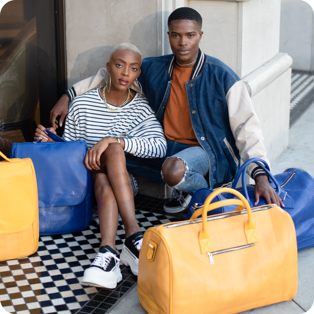 A man and a woman sit on a checkered floor entryway surrounded by blue and yellow travel bags.