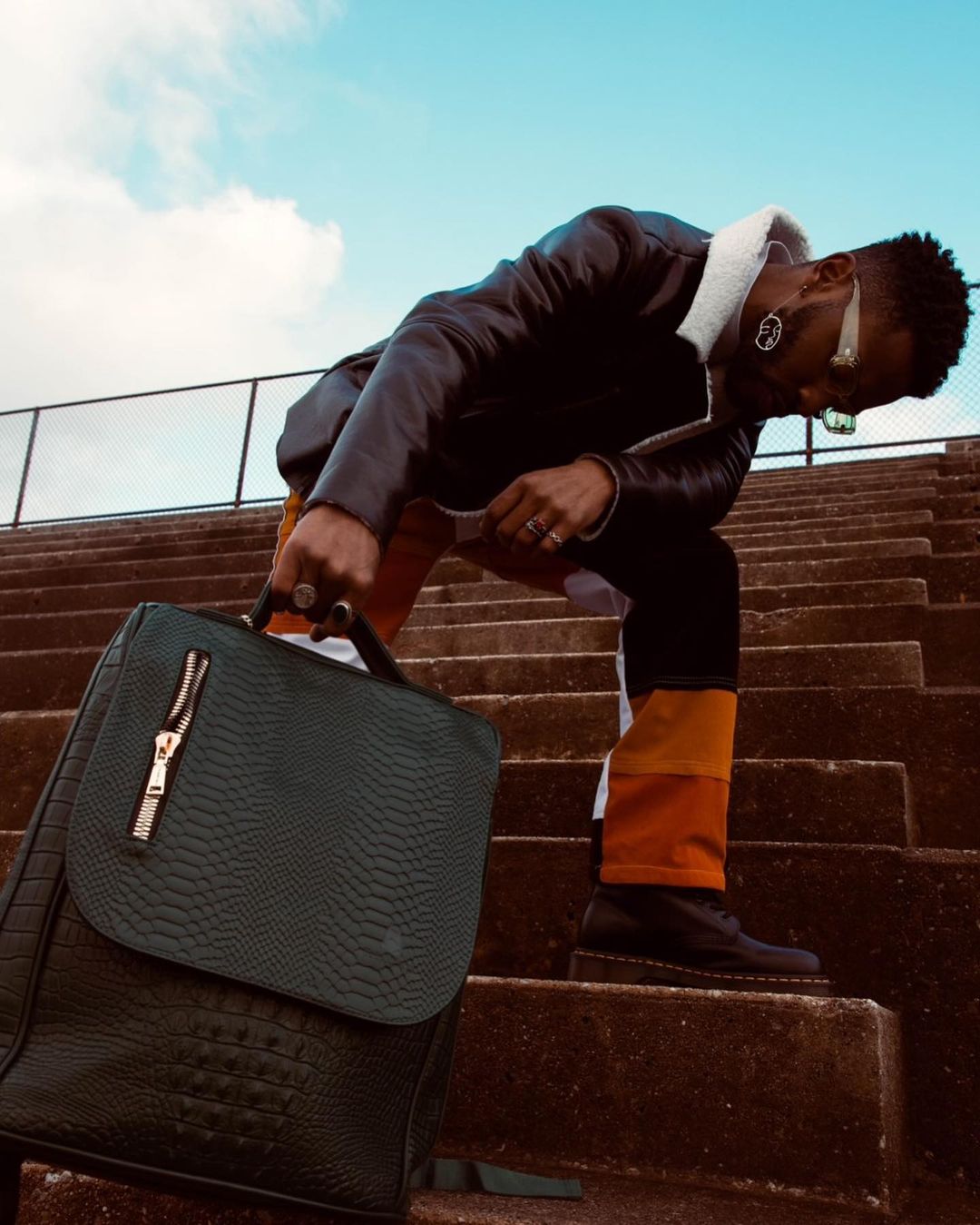 A man in a leather jacket leans over on concrete steps, holding a green textured backpack.