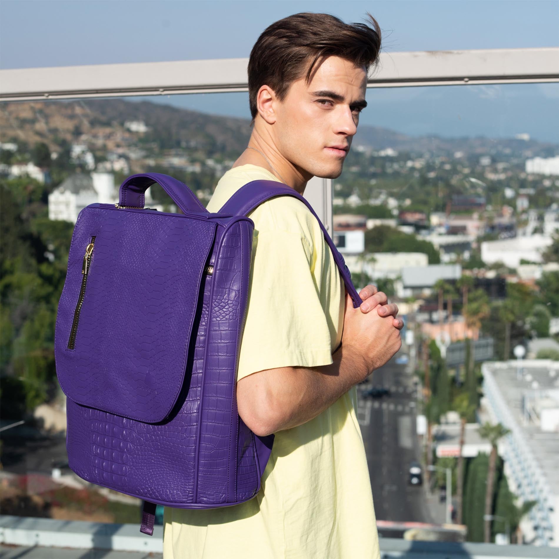 A young man in a yellow shirt carries a purple, textured backpack on a rooftop overlooking a city.