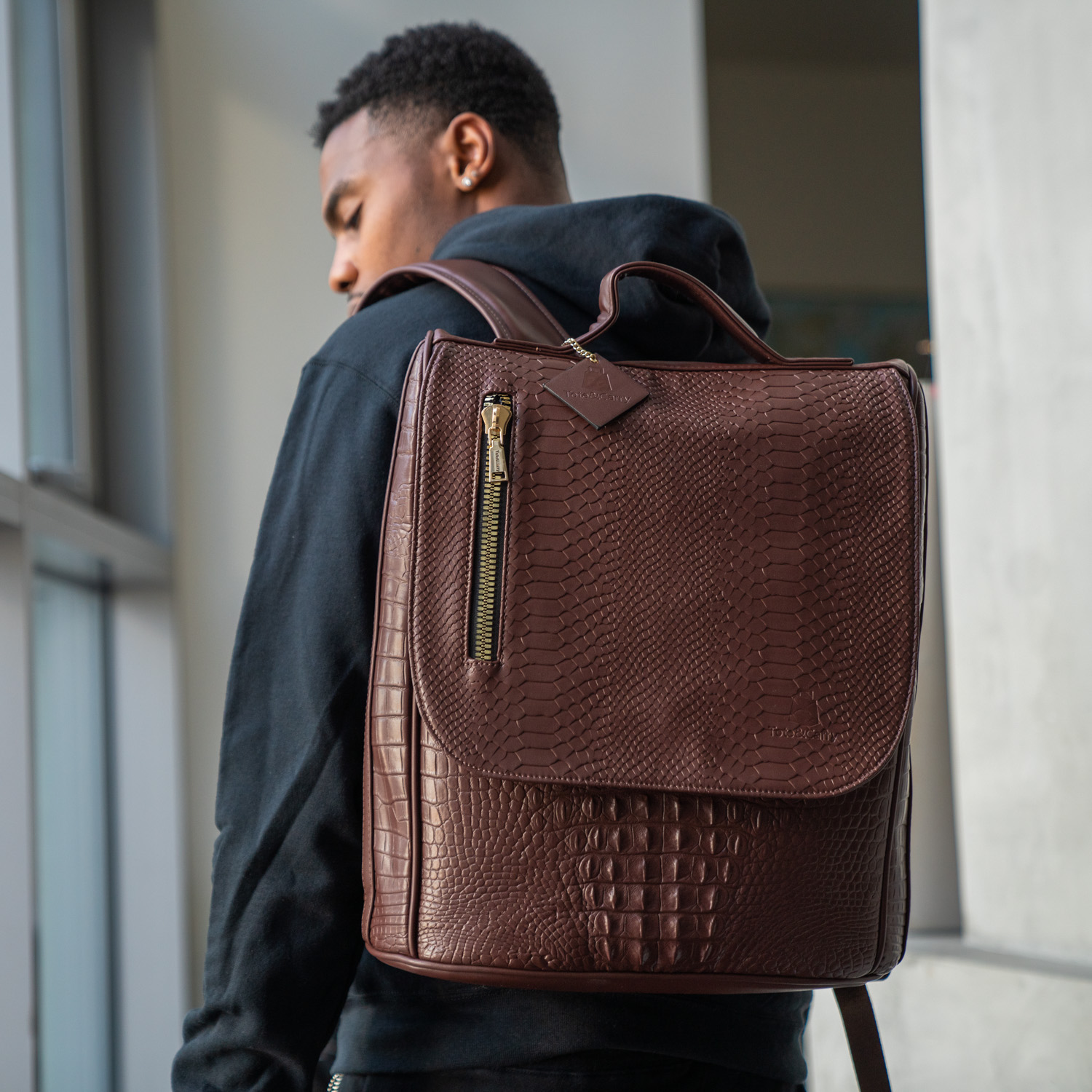 A man from behind wearing a black hoodie and a brown, textured, reptile-skin style backpack.