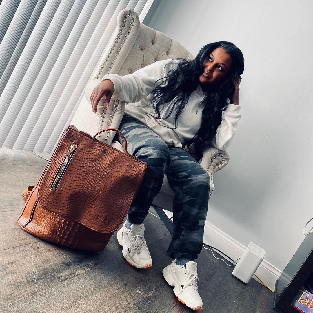 A woman in a white hoodie and camo pants sits in a high-backed chair next to a backpack.