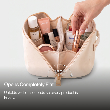 A hand placing a tube of makeup into a wide-open, cream-colored cosmetic bag filled with products.