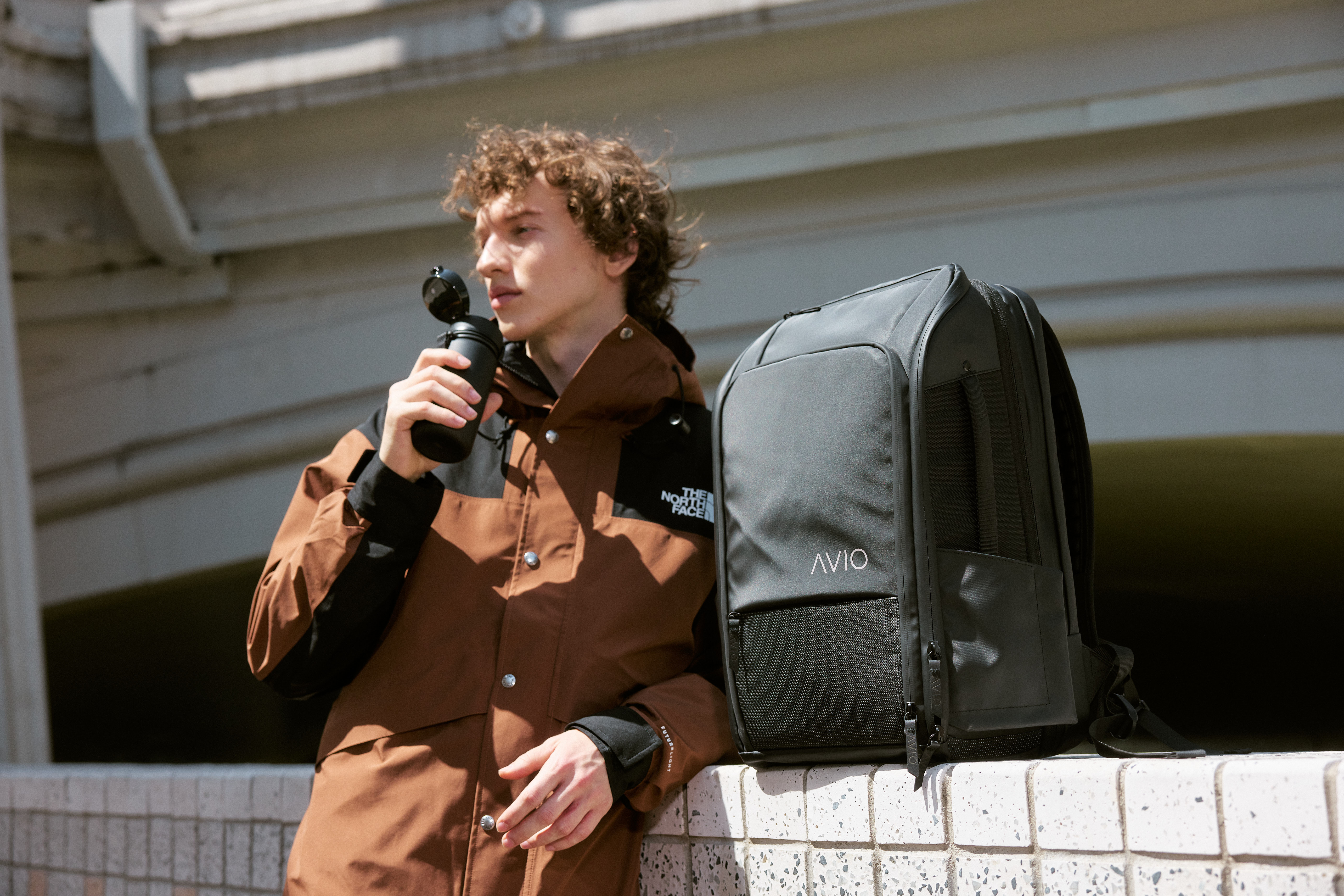 Person in brown jacket holding a bottle, with a black backpack on a ledge.