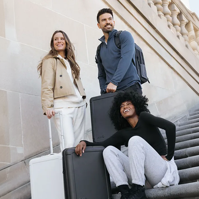 Three smiling people with their suitcases pose together on a wide set of stone stairs.
