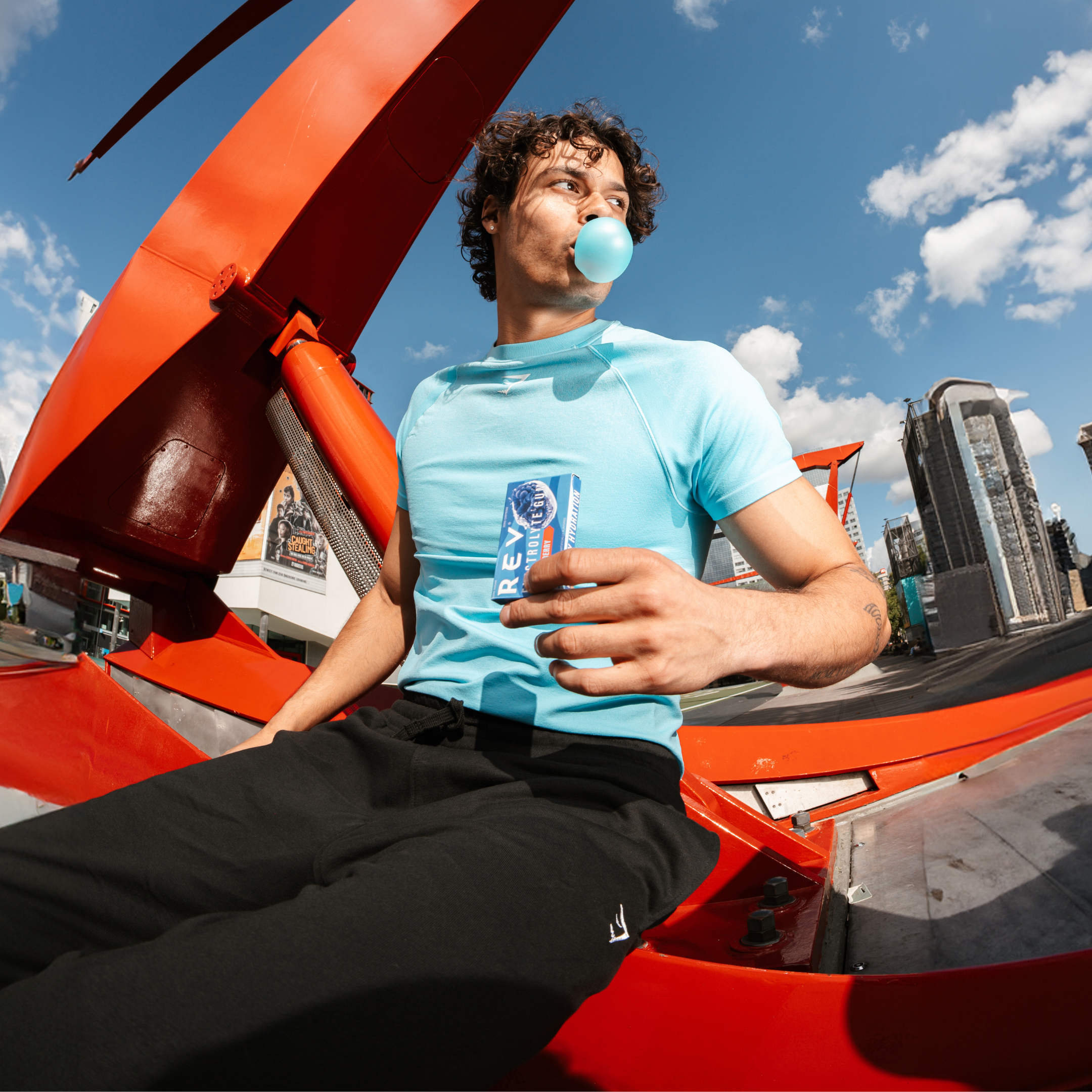 Person blowing bubble gum, holding a beverage can, with a cityscape background and large red metal structure.