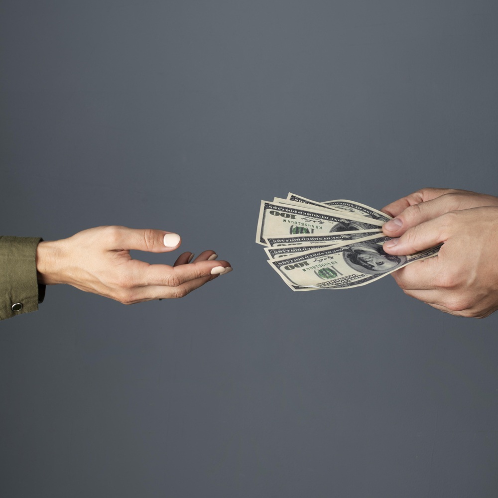 Two hands exchanging a stack of US dollar bills against a gray background.
