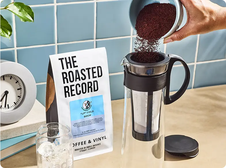 Pouring coffee grounds into a cold brew pitcher beside a coffee bag on a tiled countertop.