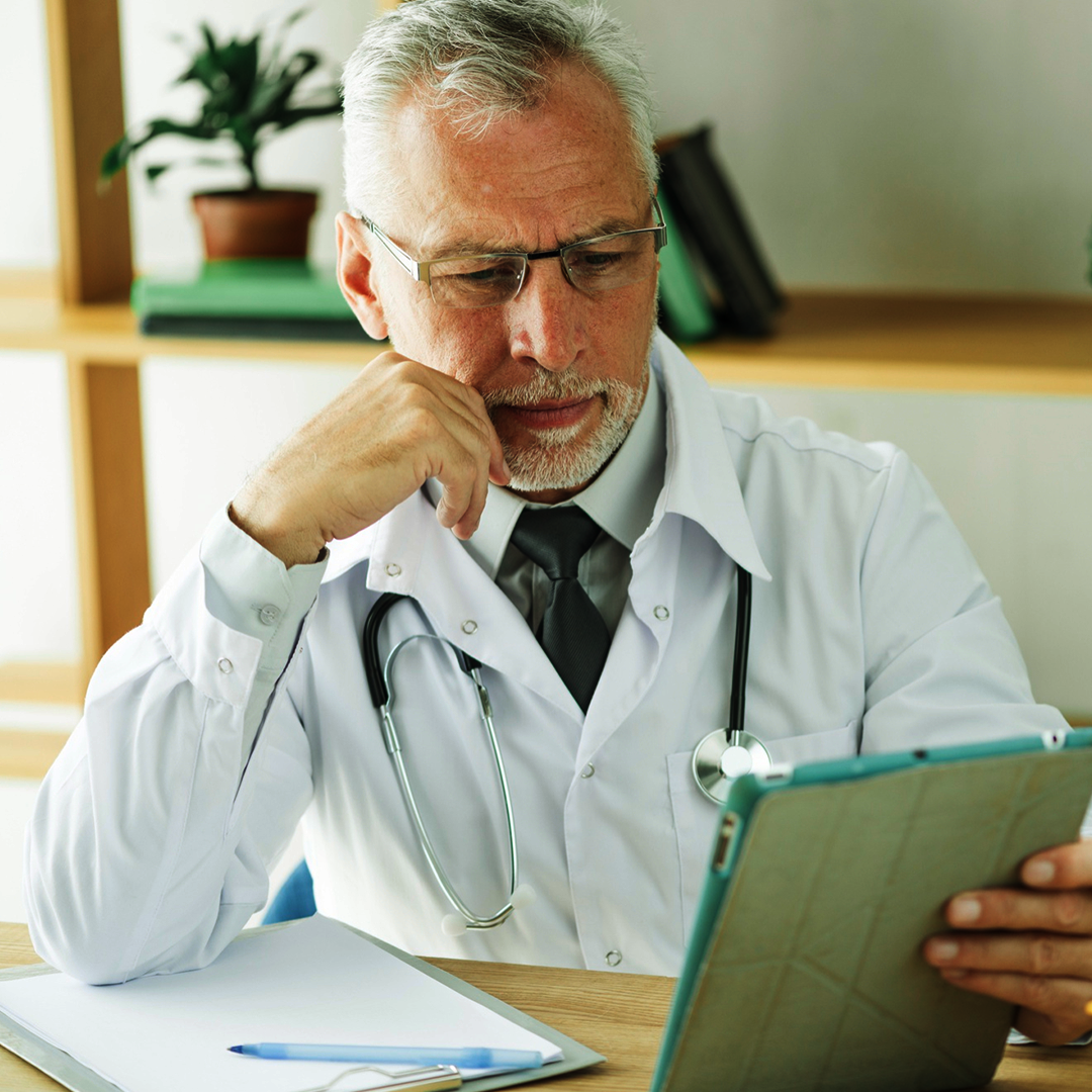 Elderly male doctor reading from a tablet, stethoscope around his neck.