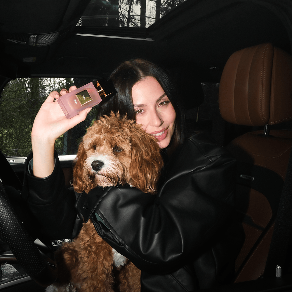A woman in a car holds a small, fluffy dog in one arm and a perfume bottle in the other.