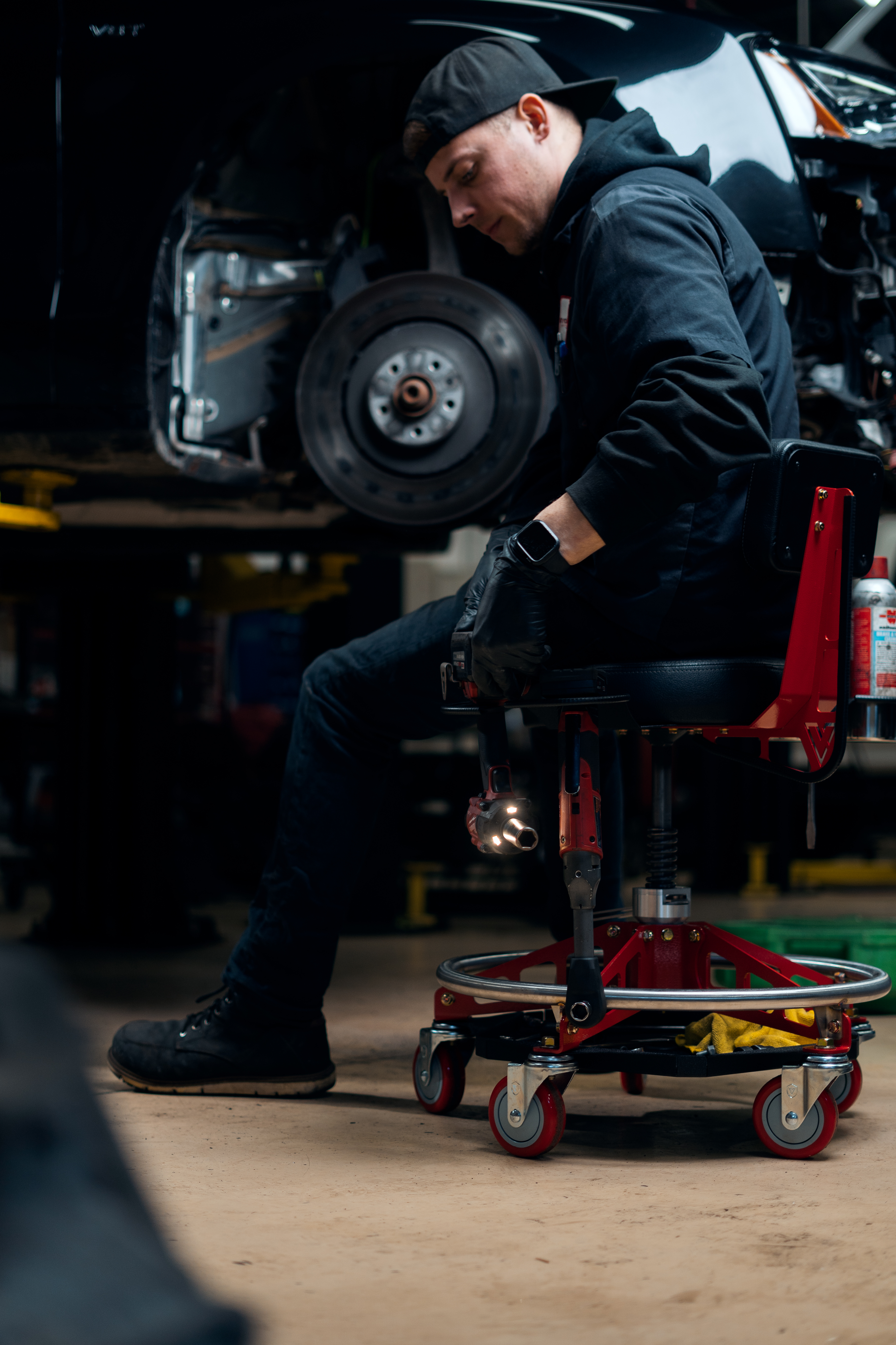 Mechanic working on a car in a garage, seated on a wheeled chair.
