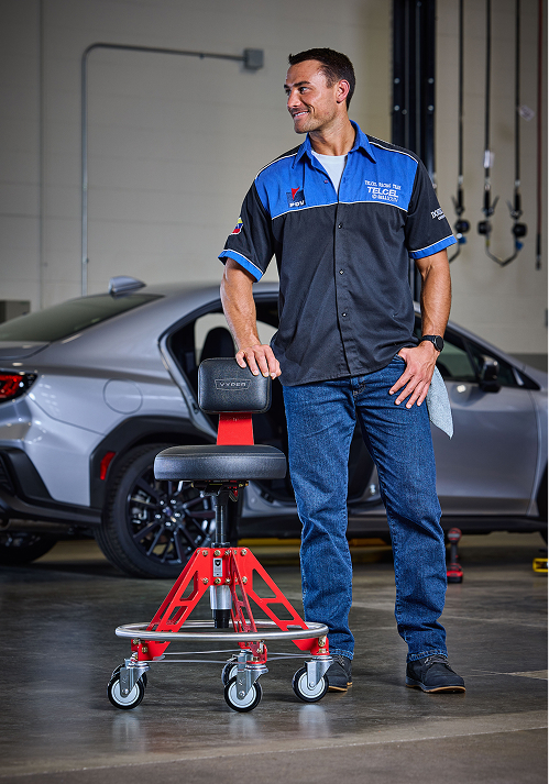 A man in a mechanic's shirt stands in a garage next to a red rolling stool and a car.