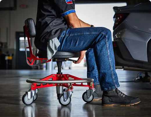 A person sits on a red rolling mechanic's stool in a garage next to a car.