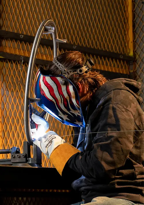 A welder wearing an American flag helmet works on a curved piece of metal.