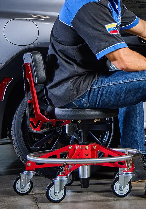 A mechanic sits on a red rolling shop stool with a backrest next to a car.